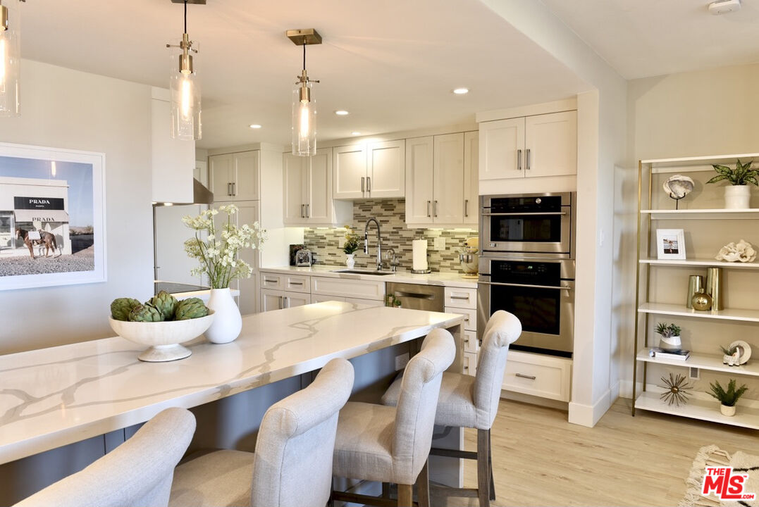 Bright staged kitchen with a marble island, white cabinetry, mosaic backsplash, and built-in double ovens.