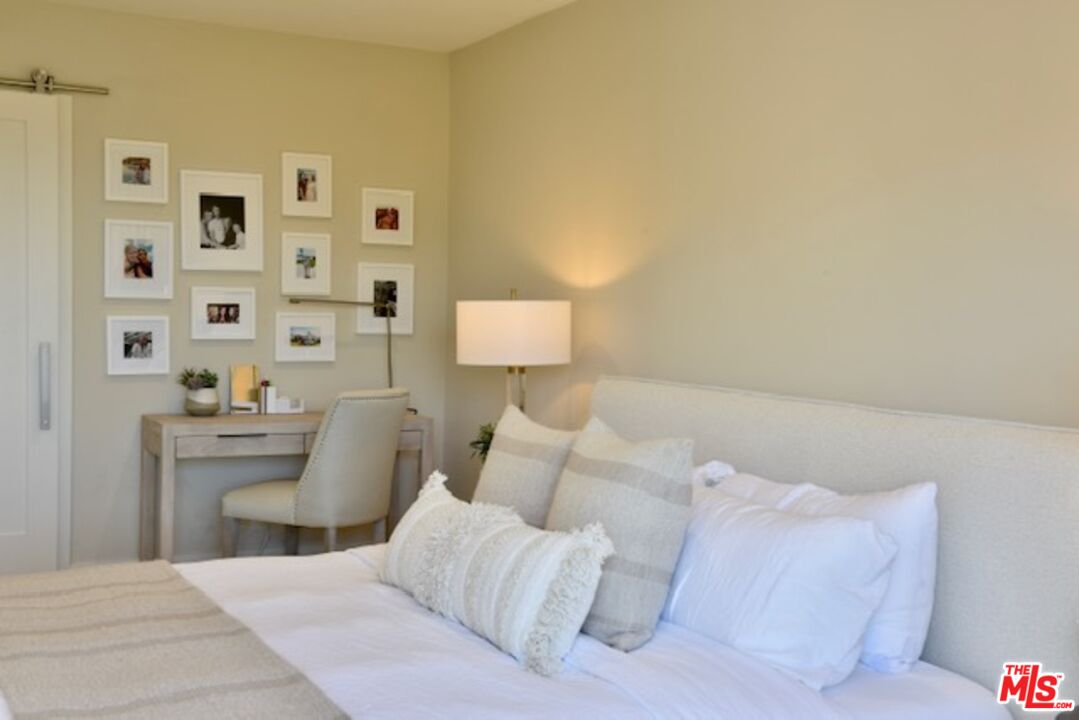 Bedroom with a neatly made bed, framed photos, and a small desk beside the closet.