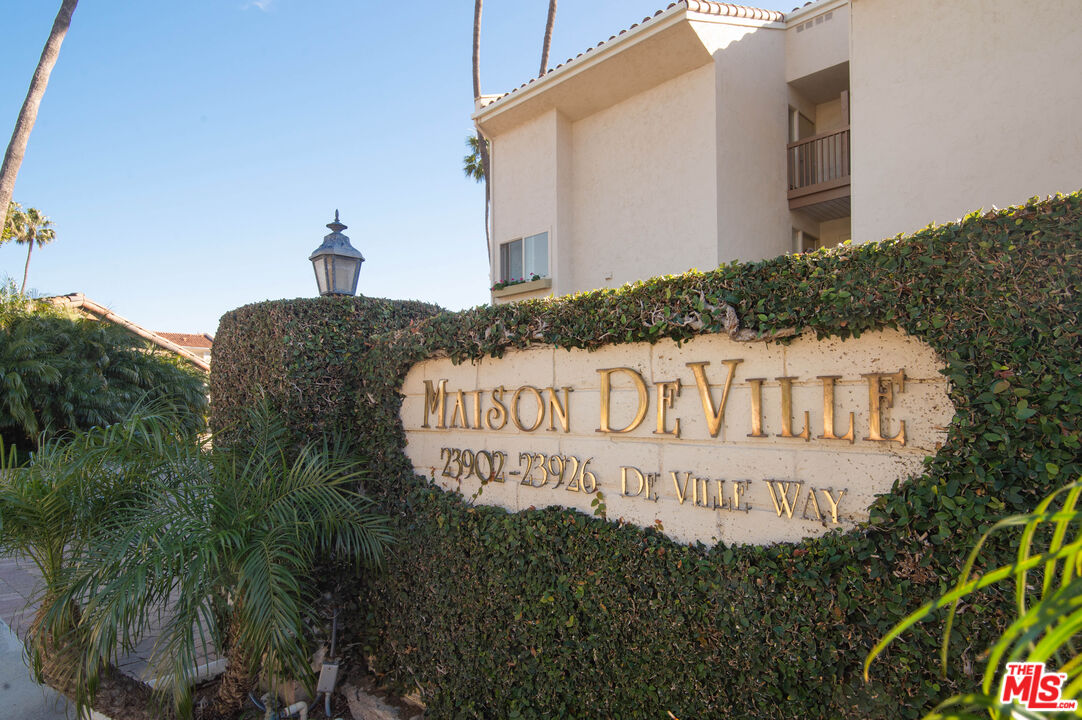 Maison De Ville entrance sign beside a cream stucco condominium building and tropical landscaping.