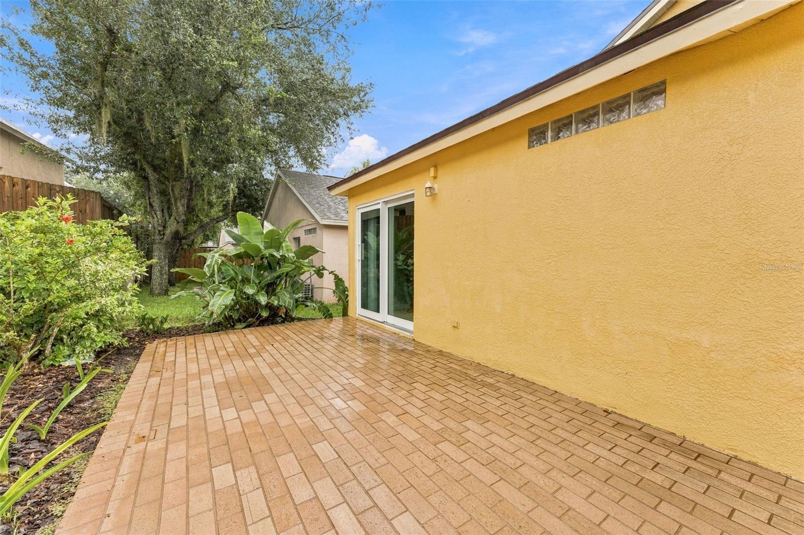 Brick-paved patio beside a yellow stucco home, with sliding glass doors and tropical landscaping.