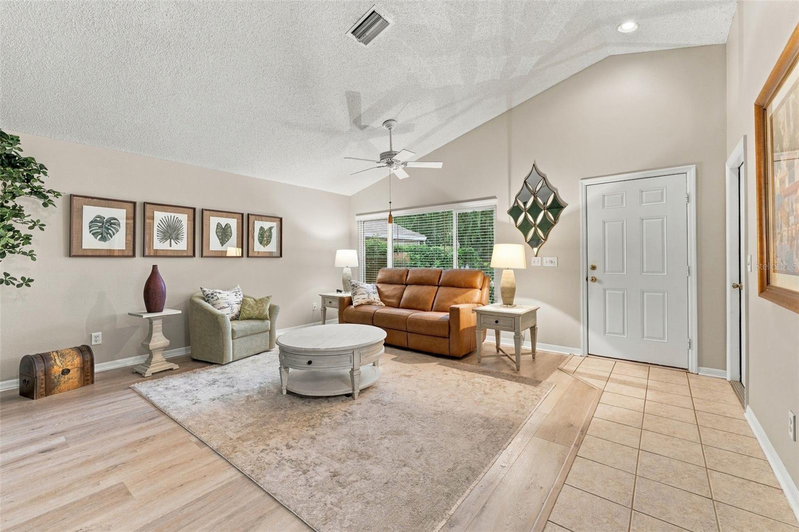 Furnished living room with vaulted ceiling, leather sofa, and large window overlooking greenery.