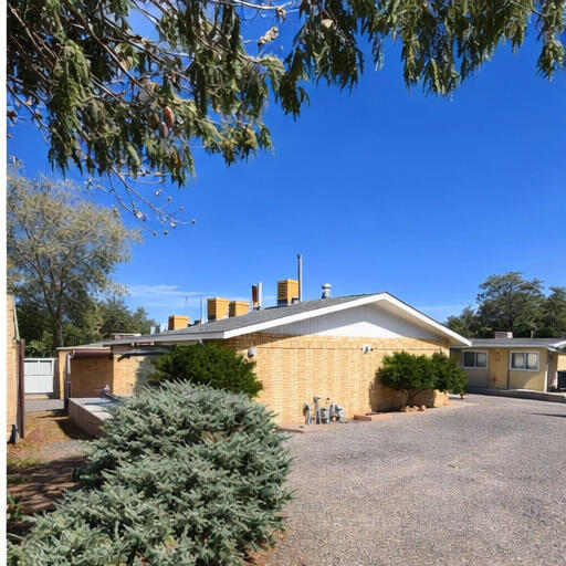 Single-story brick multi-family building with a gravel parking area and mature trees.