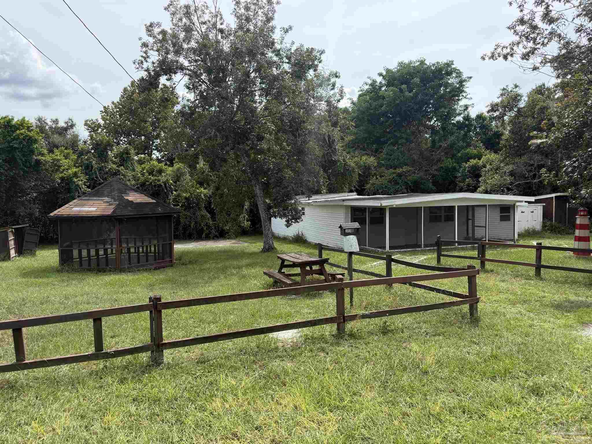 Manufactured home behind a fenced lawn, with a screened gazebo and mature trees.