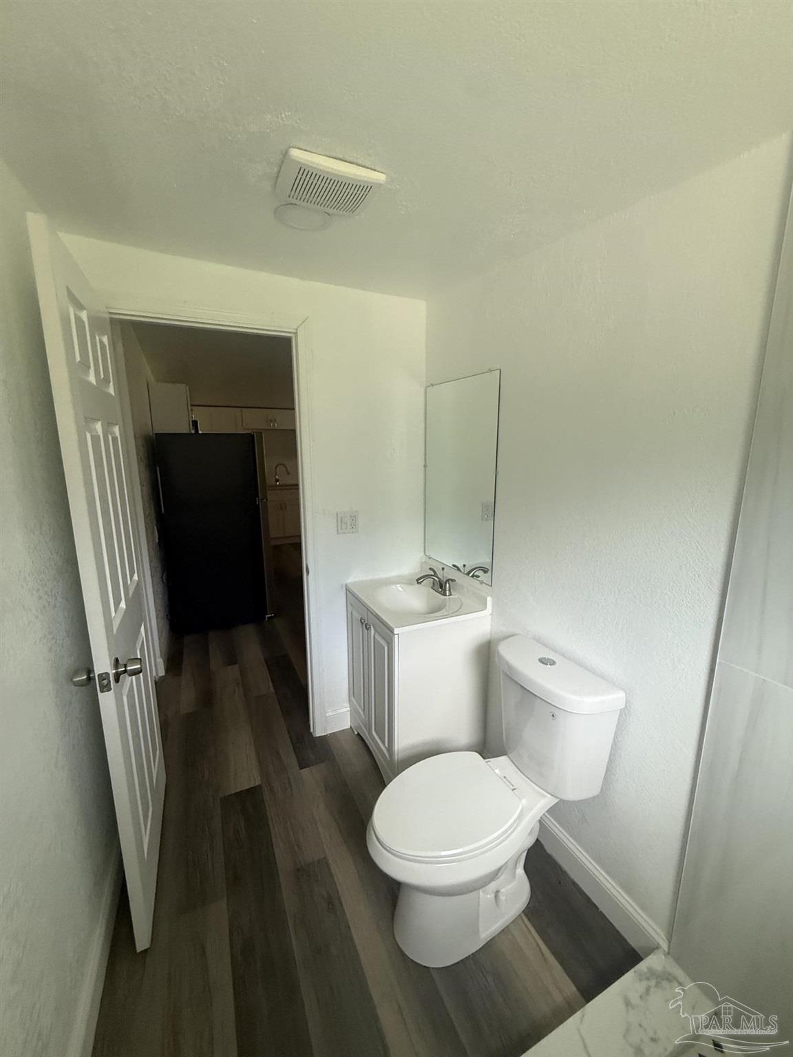Bathroom with a white toilet and vanity, mirror, and dark plank flooring; kitchen visible through the open doorway.