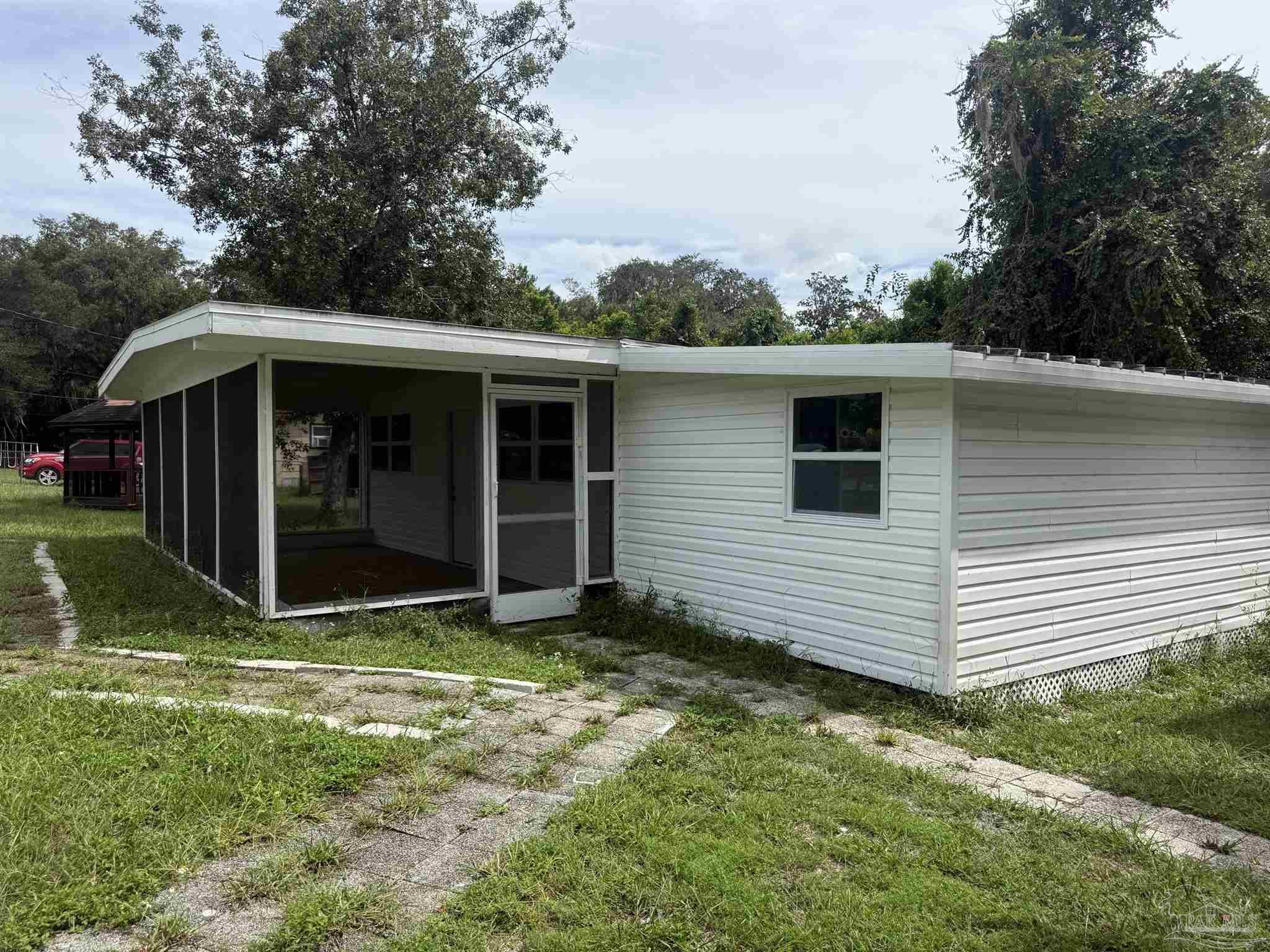 White manufactured home with a screened porch, overgrown walkway, and mature trees.