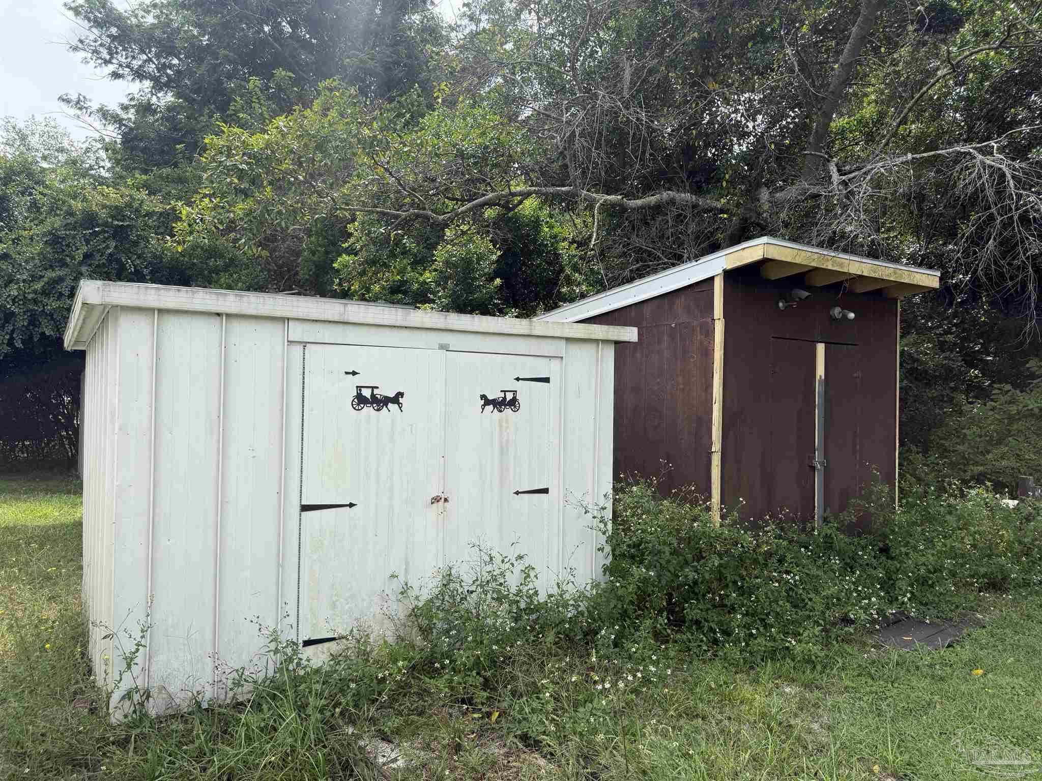 Two storage sheds among trees, with a white decorated shed in front of a dark brown shed.