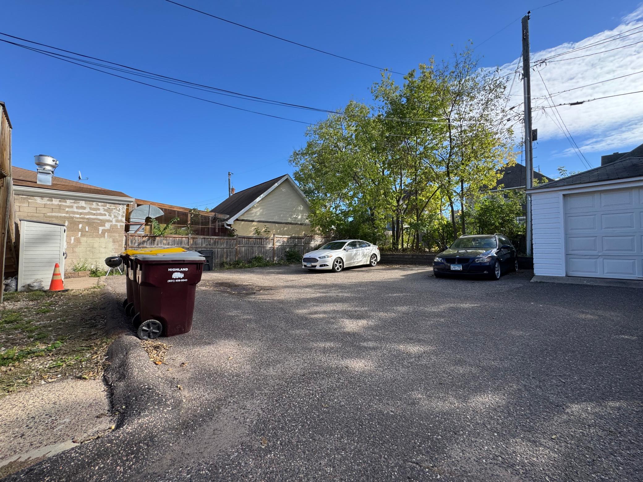 Paved parking area behind the property with two cars, a white garage, and a leafy tree.