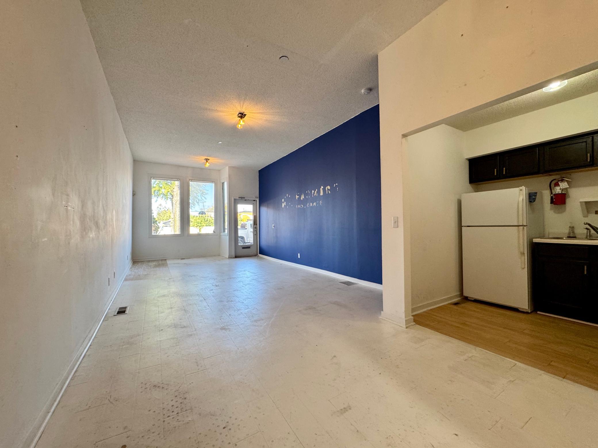 Vacant open interior with a blue accent wall, front windows and a small kitchenette.