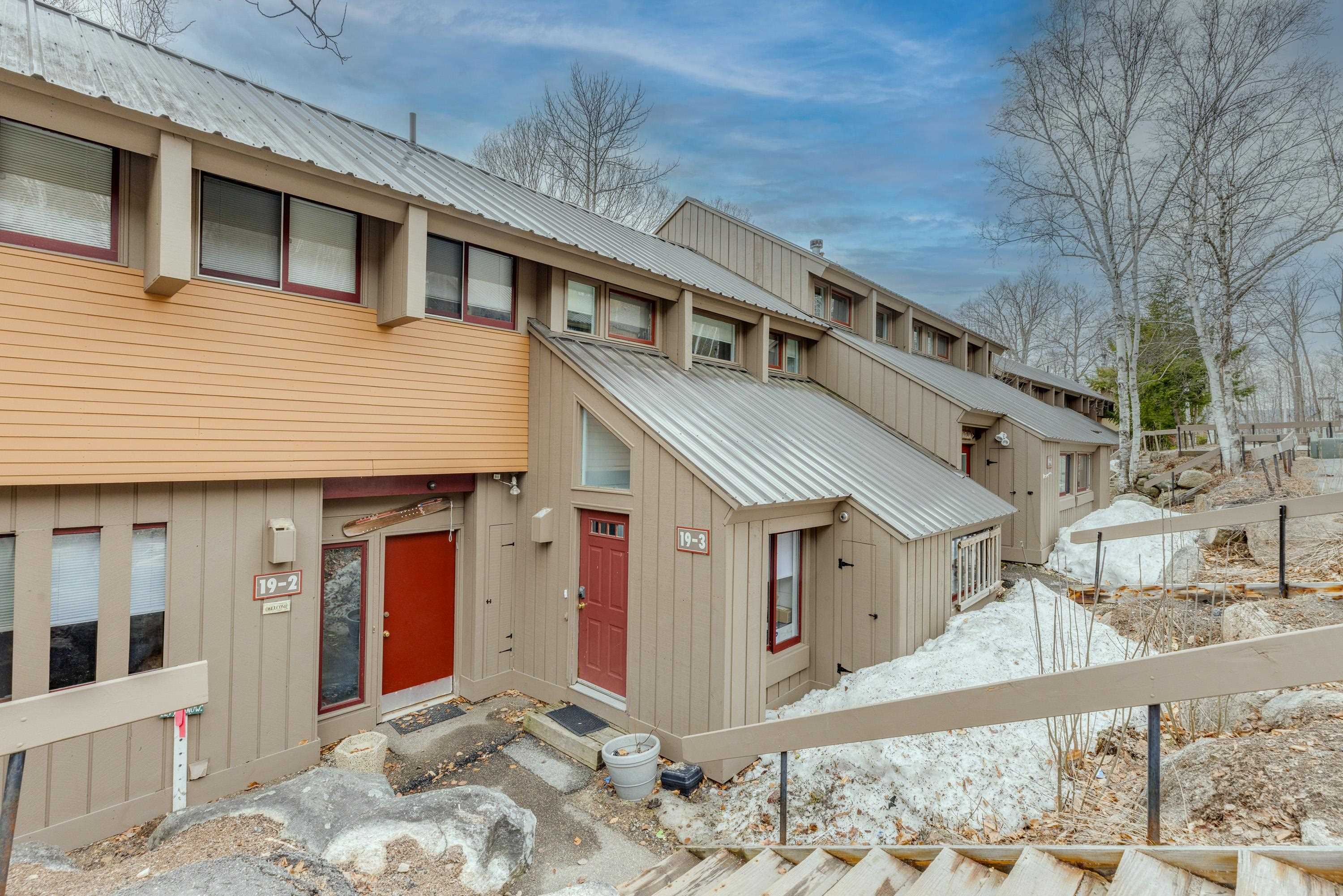 Townhouse-style condo exterior with a silver metal roof, red unit doors, and snow along the walkway.