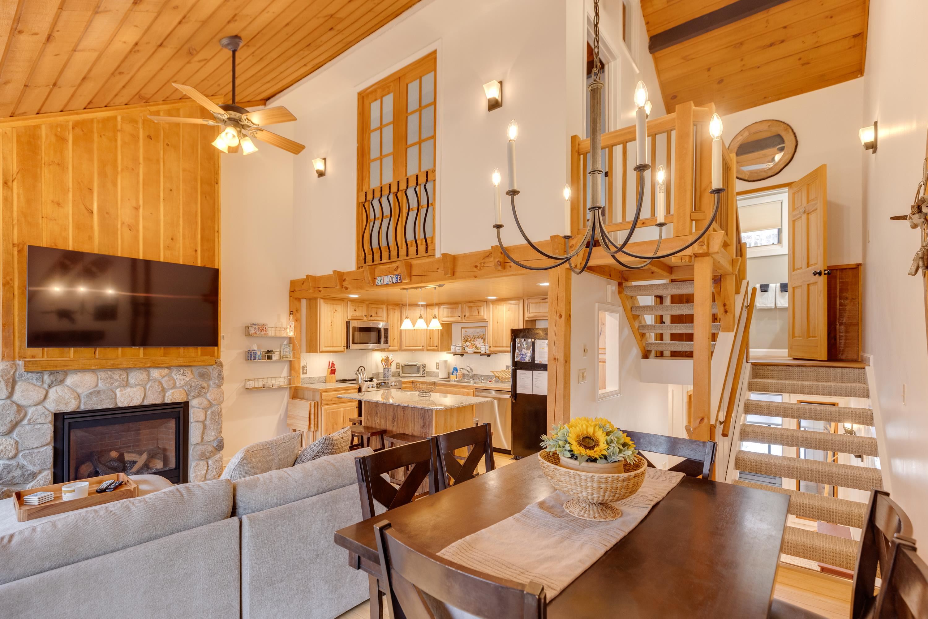 Open-plan condo living and dining area with vaulted wood ceiling, stone fireplace, kitchen, and loft staircase.