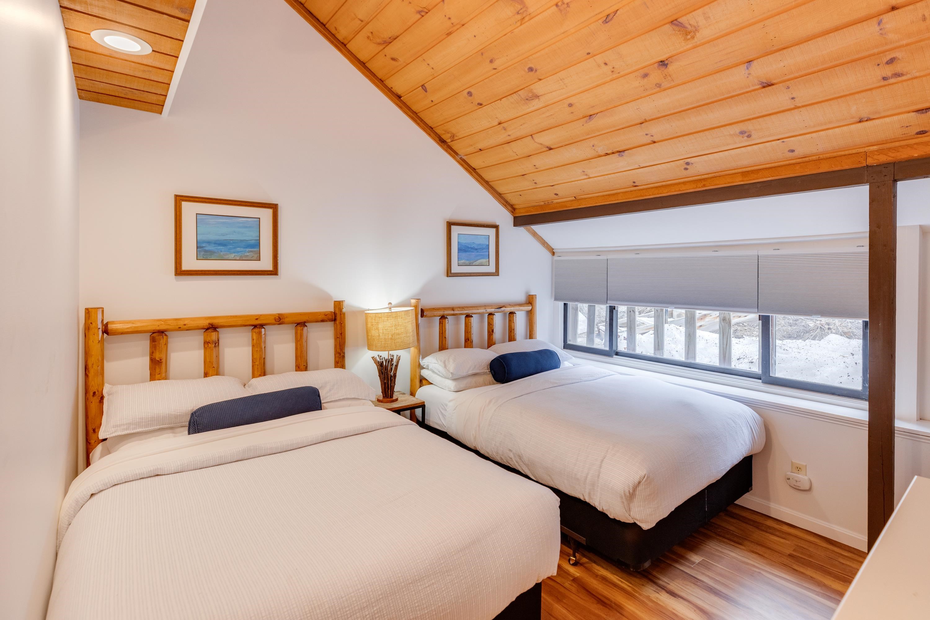 Bedroom with two white beds, rustic log headboards, and a vaulted wood ceiling beside a snowy window.