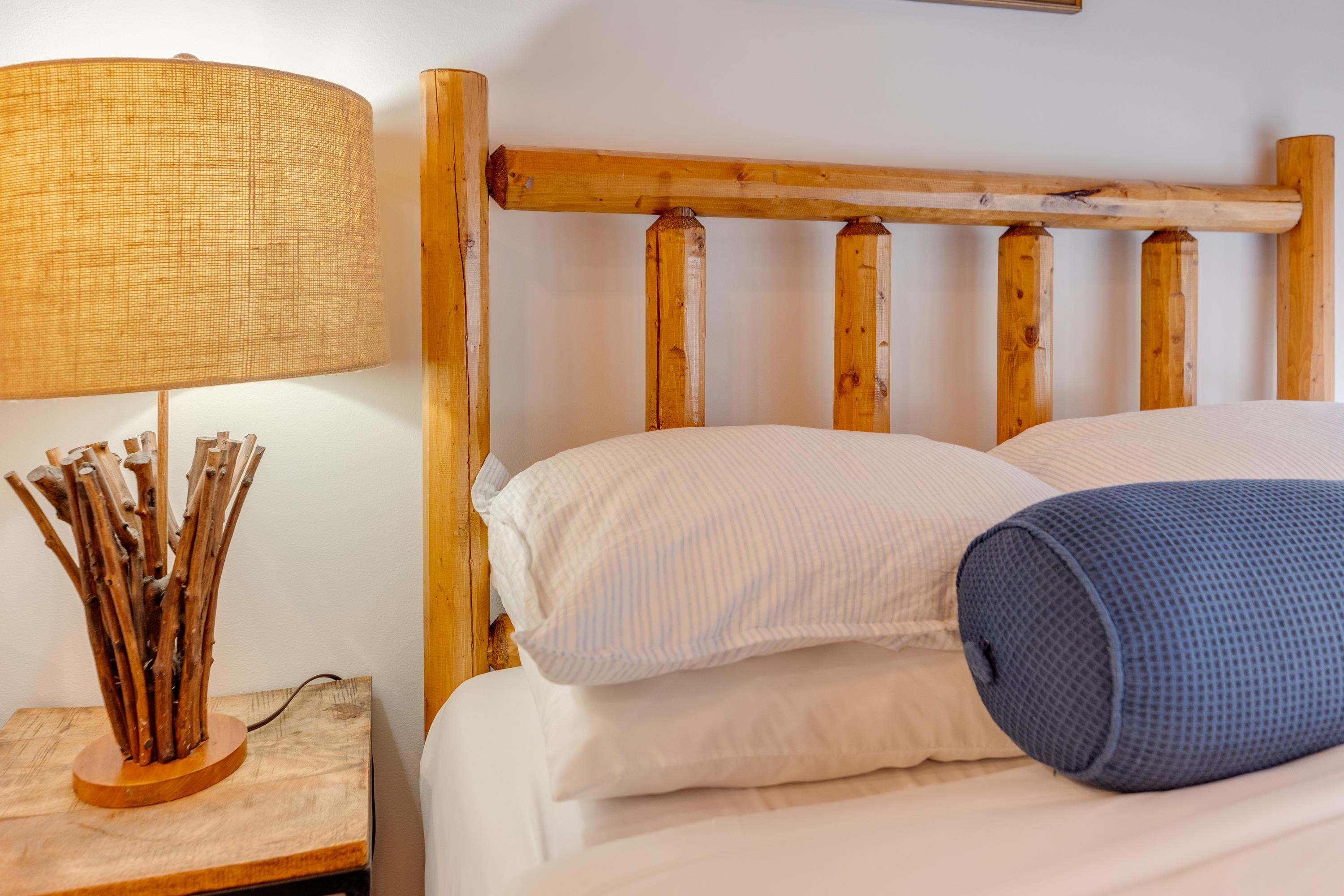 Bedroom with rustic wooden bed frame, white striped bedding, blue bolster pillow, and woven-shade bedside lamp.