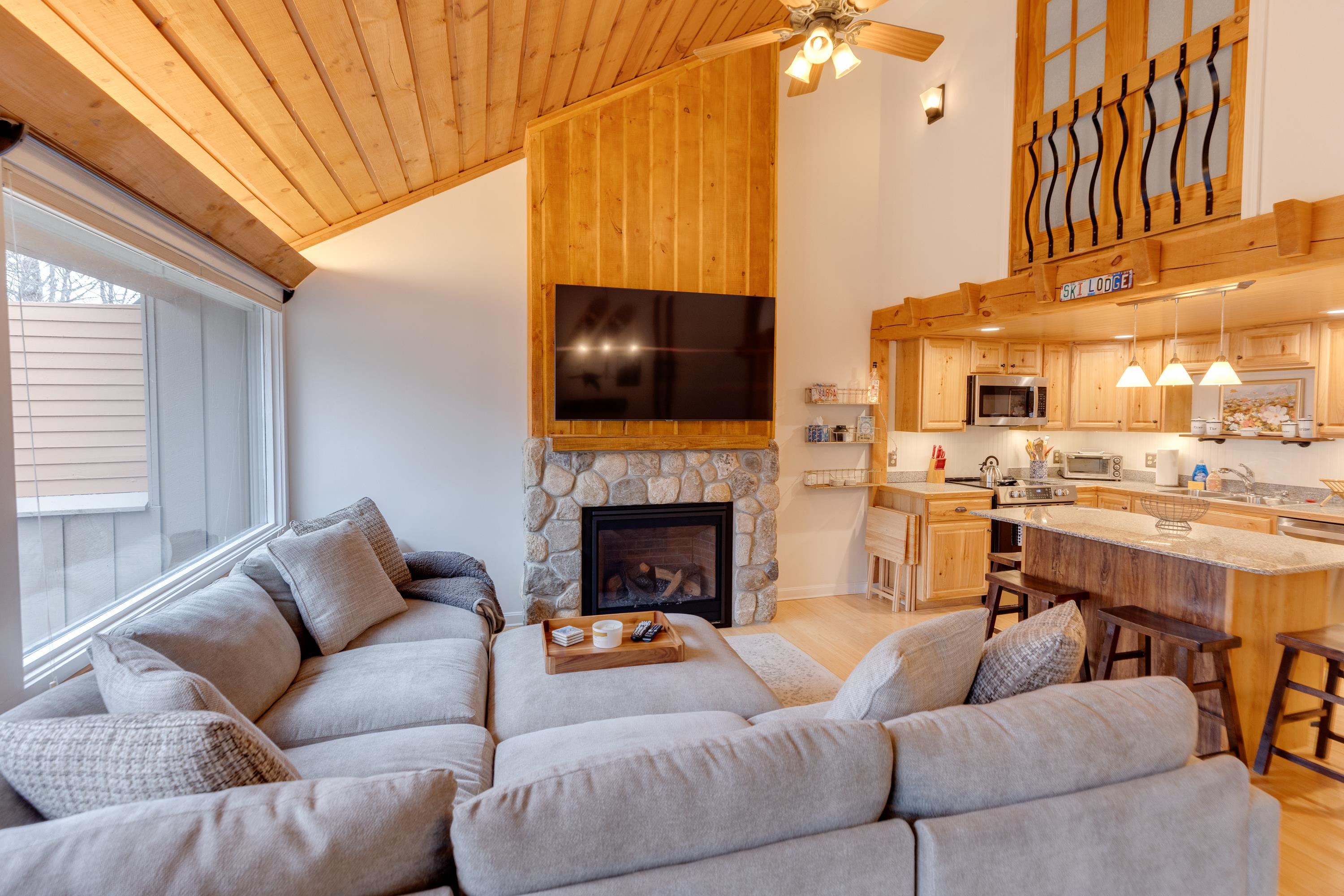 Living room with a gray sectional facing a stone fireplace, beneath a vaulted wood ceiling beside an open kitchen.