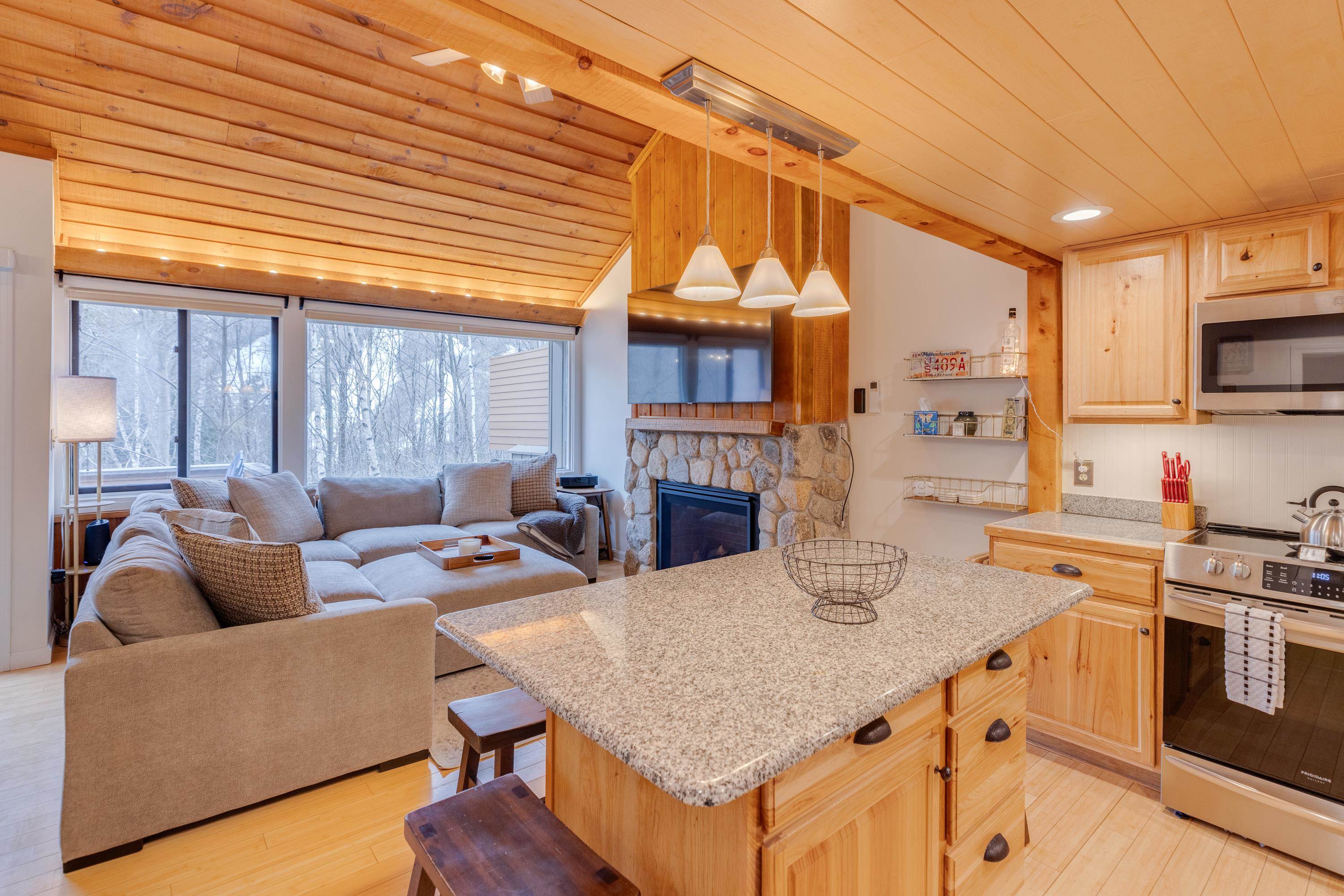 Open-plan kitchen and living room with a granite island, stone fireplace, and large windows overlooking winter woods.