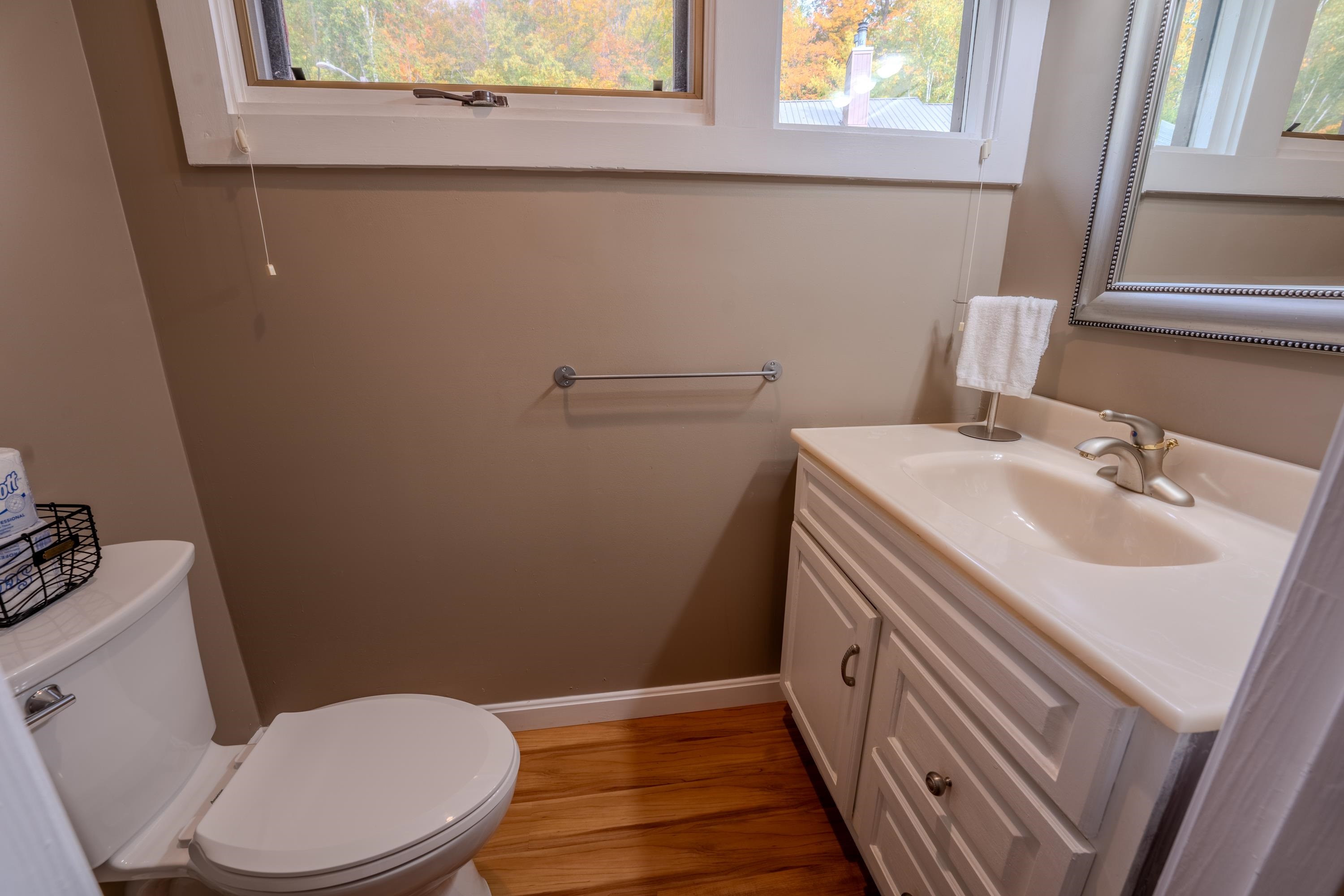 Bathroom with a white toilet, paneled vanity, and high windows overlooking autumn foliage.