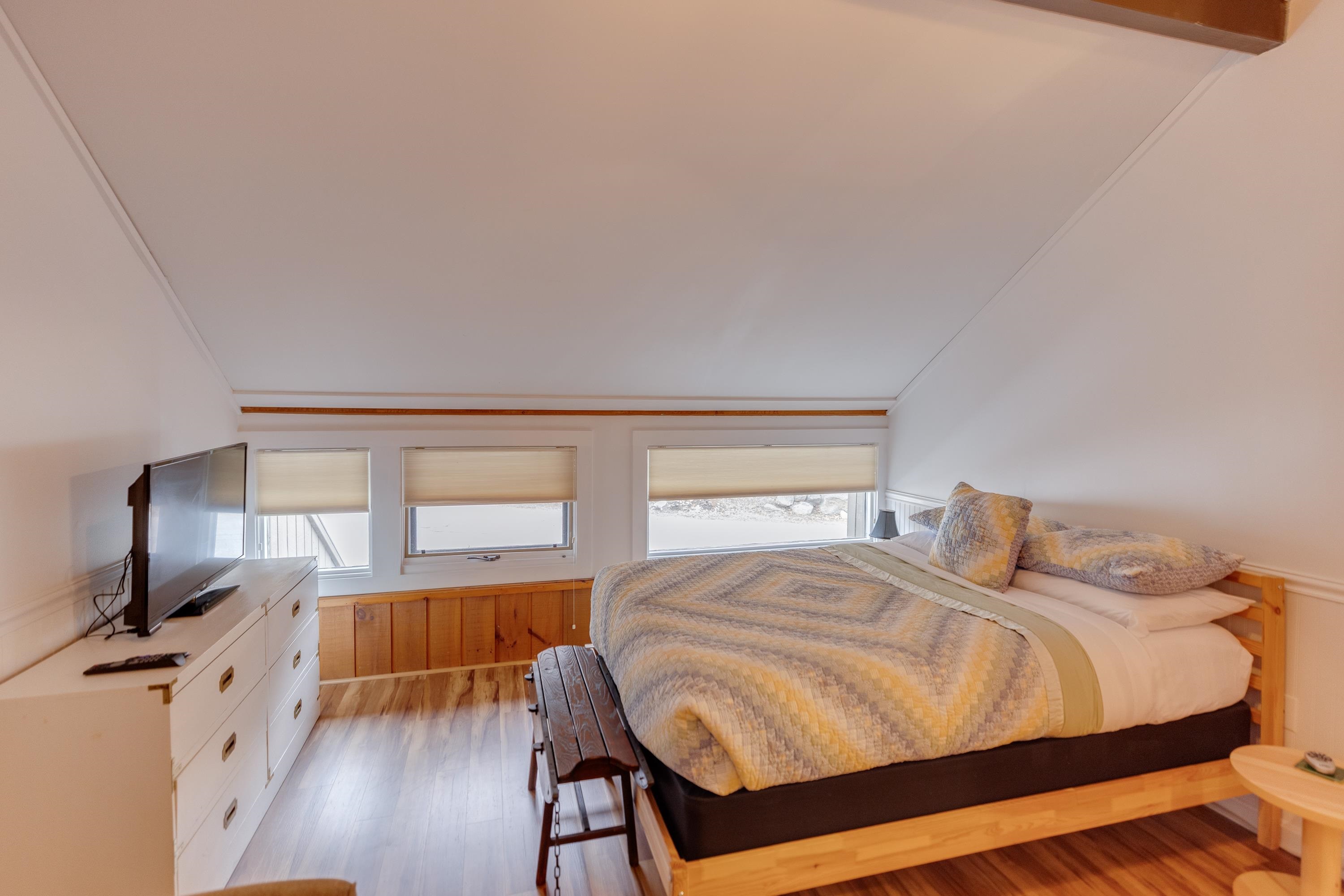 Furnished bedroom with a sloped ceiling, large windows, and a wooden bed with a patterned quilt.