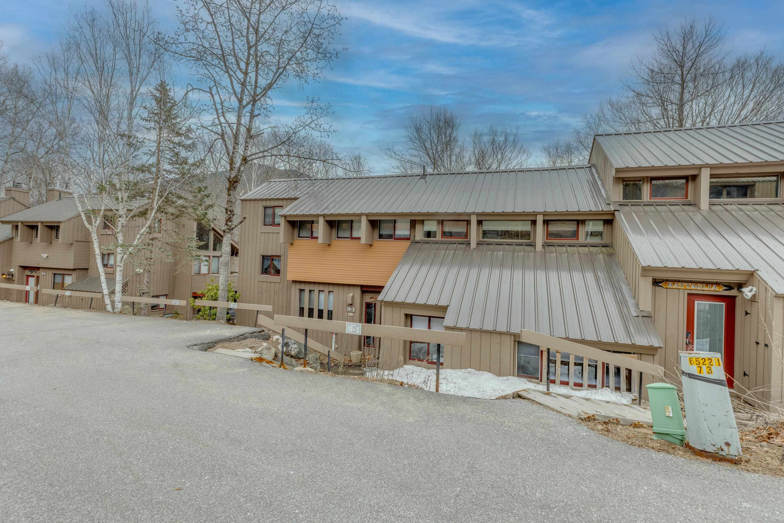 Multi-unit building with tan siding, gray metal roofs, and patches of snow beside the driveway.