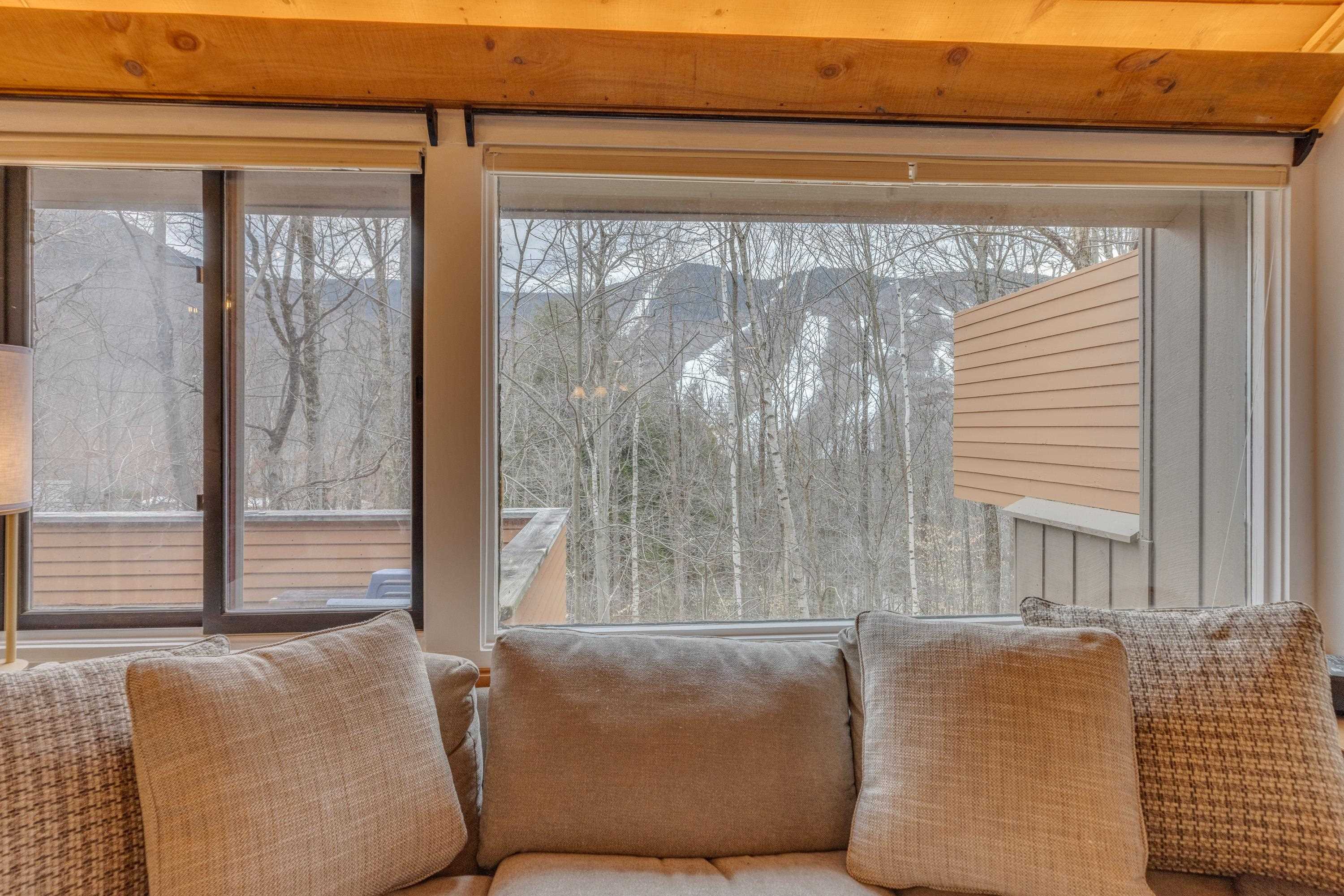 Living room sofa beneath wide windows overlooking leafless woods, with exposed wood ceiling beams.