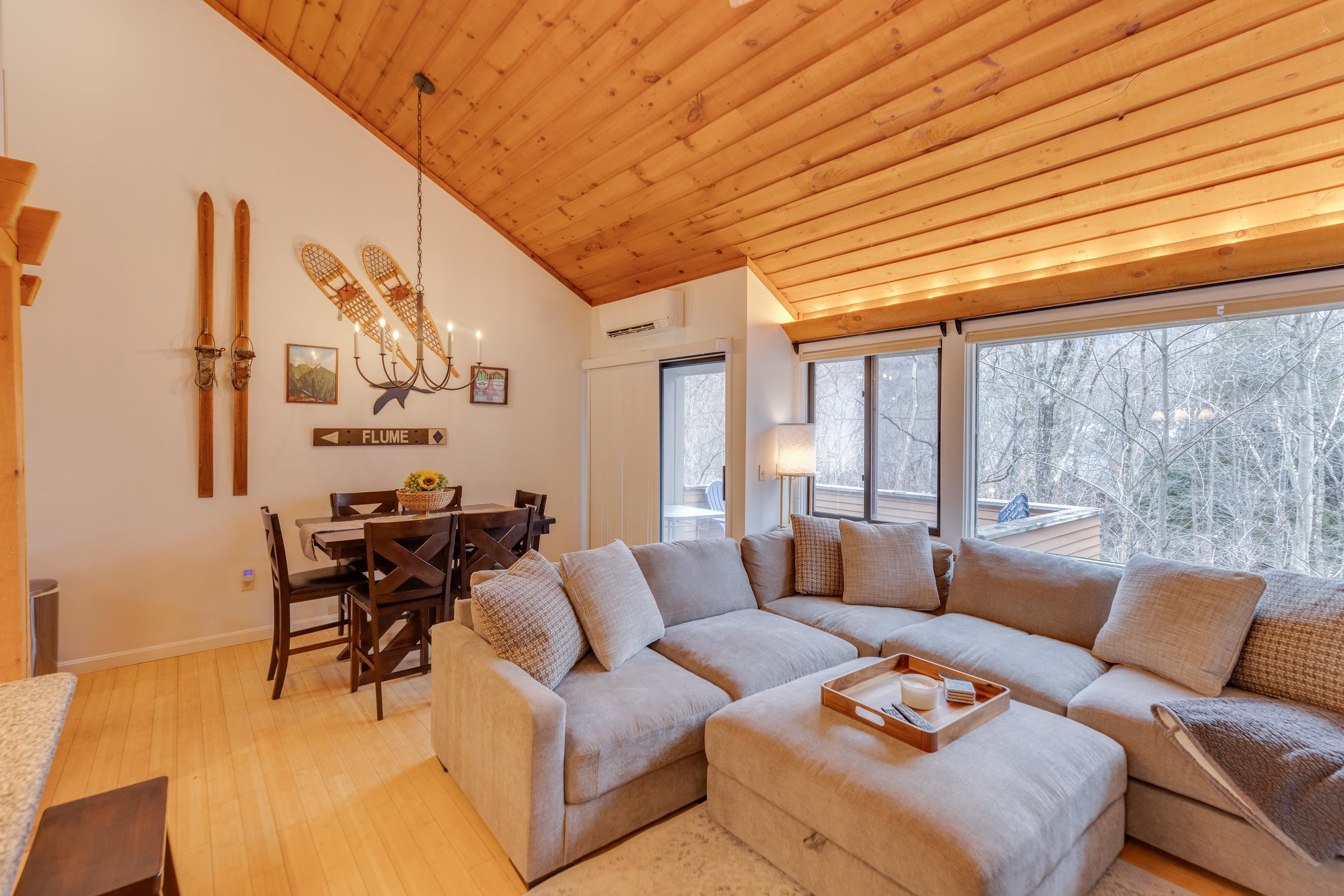 Living room with a vaulted wood ceiling, gray sectional, dining area, and windows overlooking snowy woods.