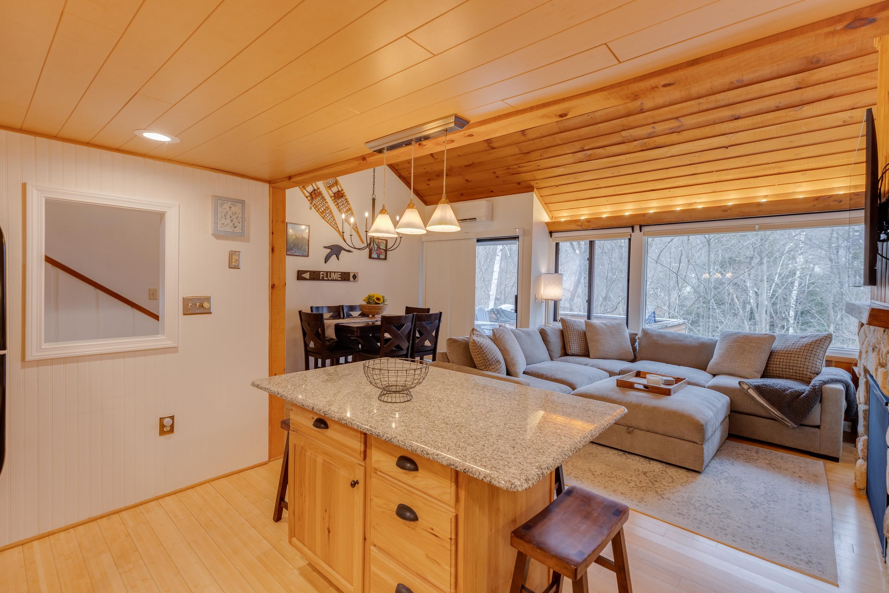 Open-plan living and dining area with a granite island, vaulted wood ceiling, sectional sofa, and large windows.