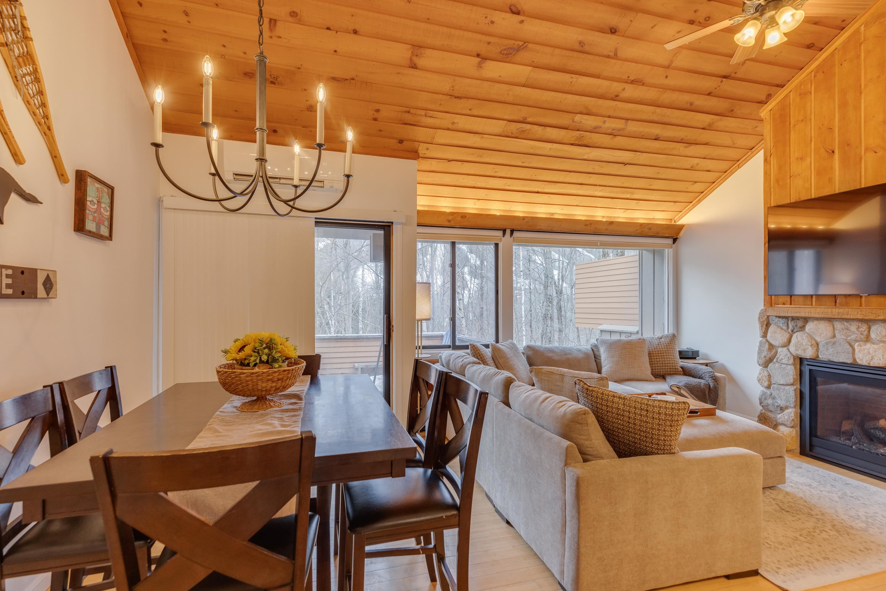 Open-plan living and dining room with a vaulted wood ceiling, stone fireplace, and windows overlooking winter trees.