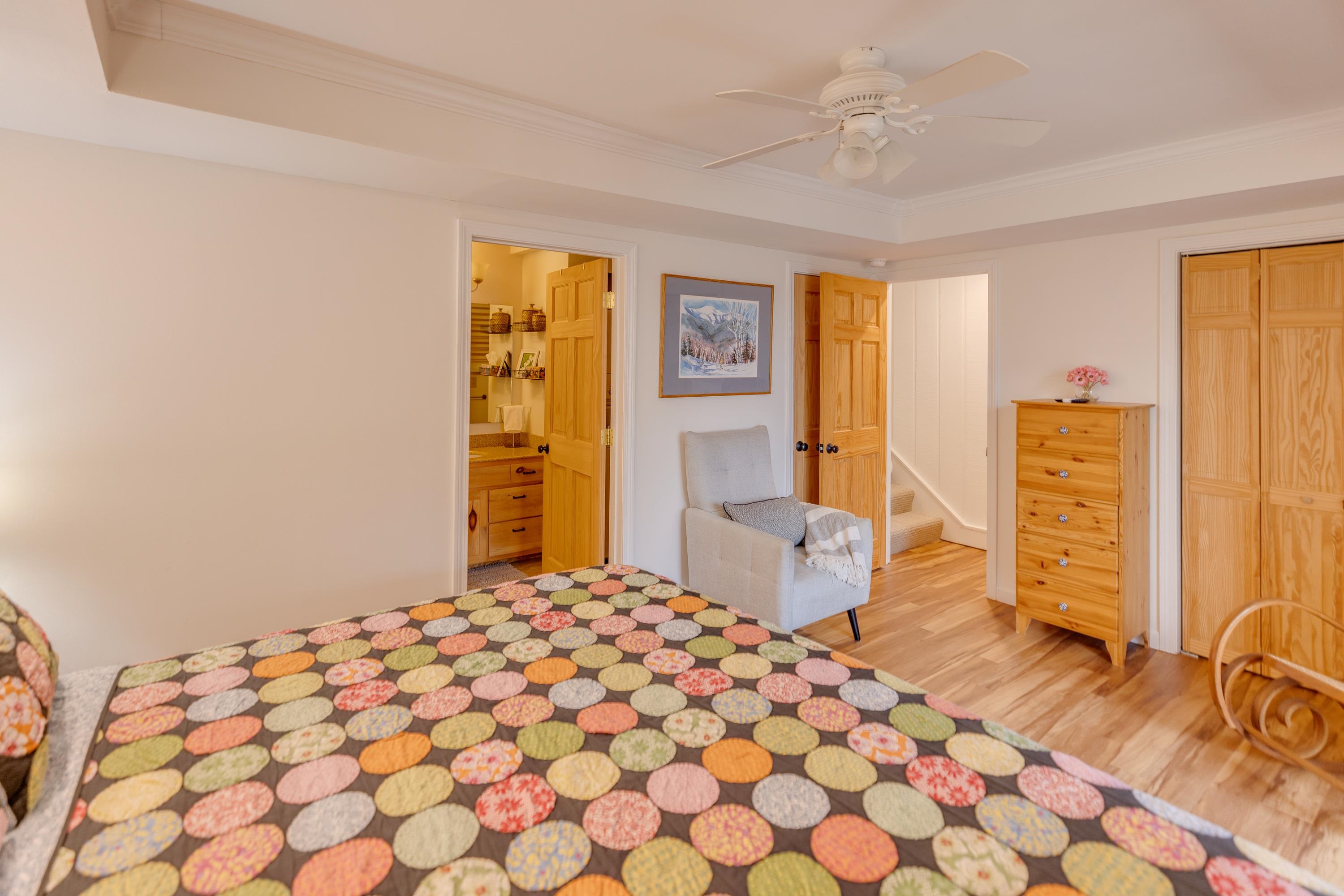 Bedroom with a colorful patchwork quilt, pine furniture, and open doorways to a bathroom and staircase.