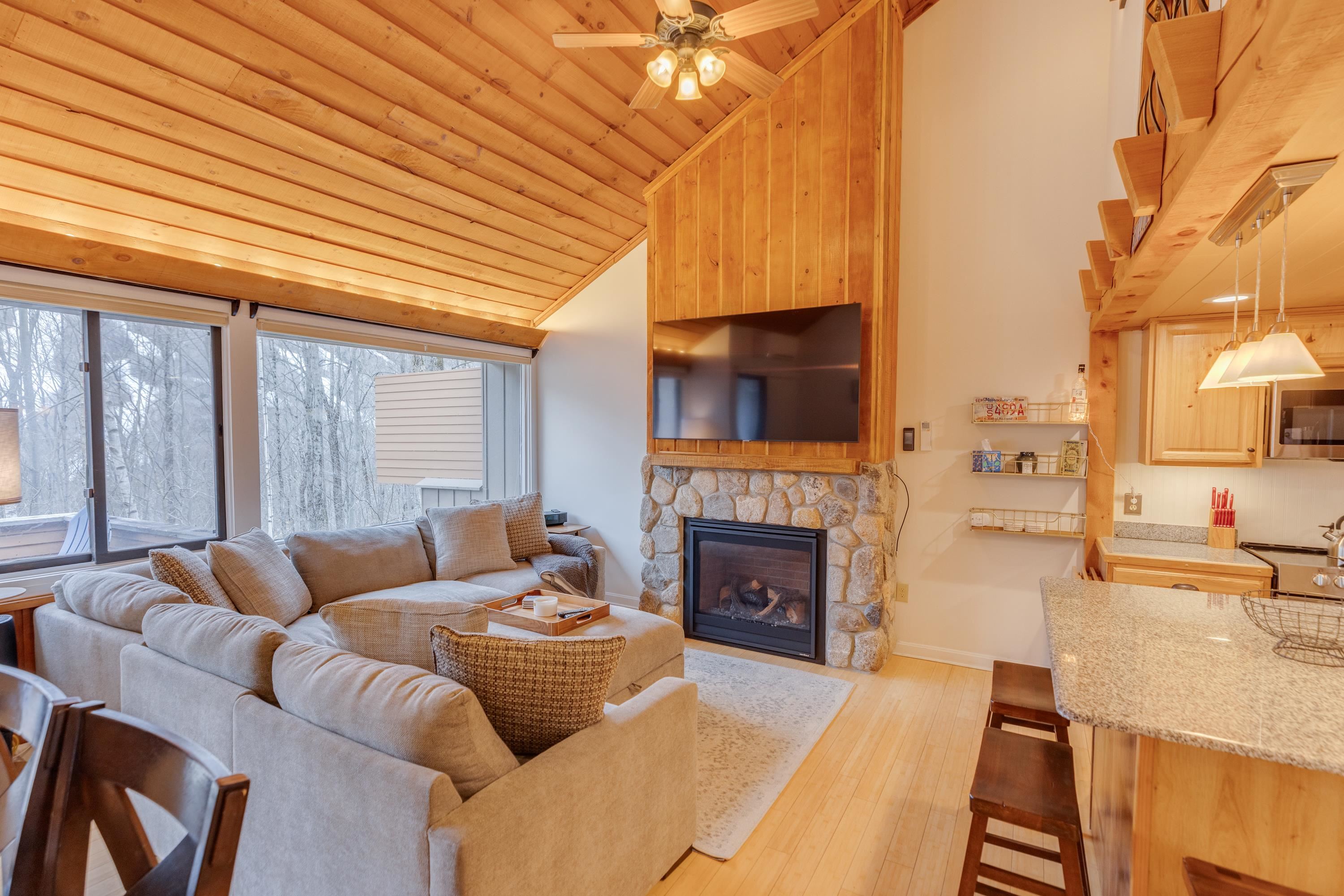 Living room with a vaulted wood ceiling, stone fireplace, and large windows overlooking snowy trees.