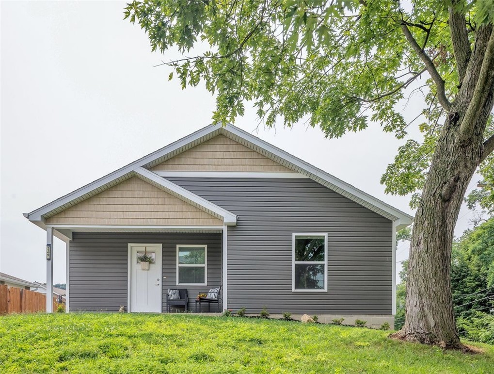 Gray gabled house with a small covered porch and chairs, set behind a grassy lawn and large tree.
