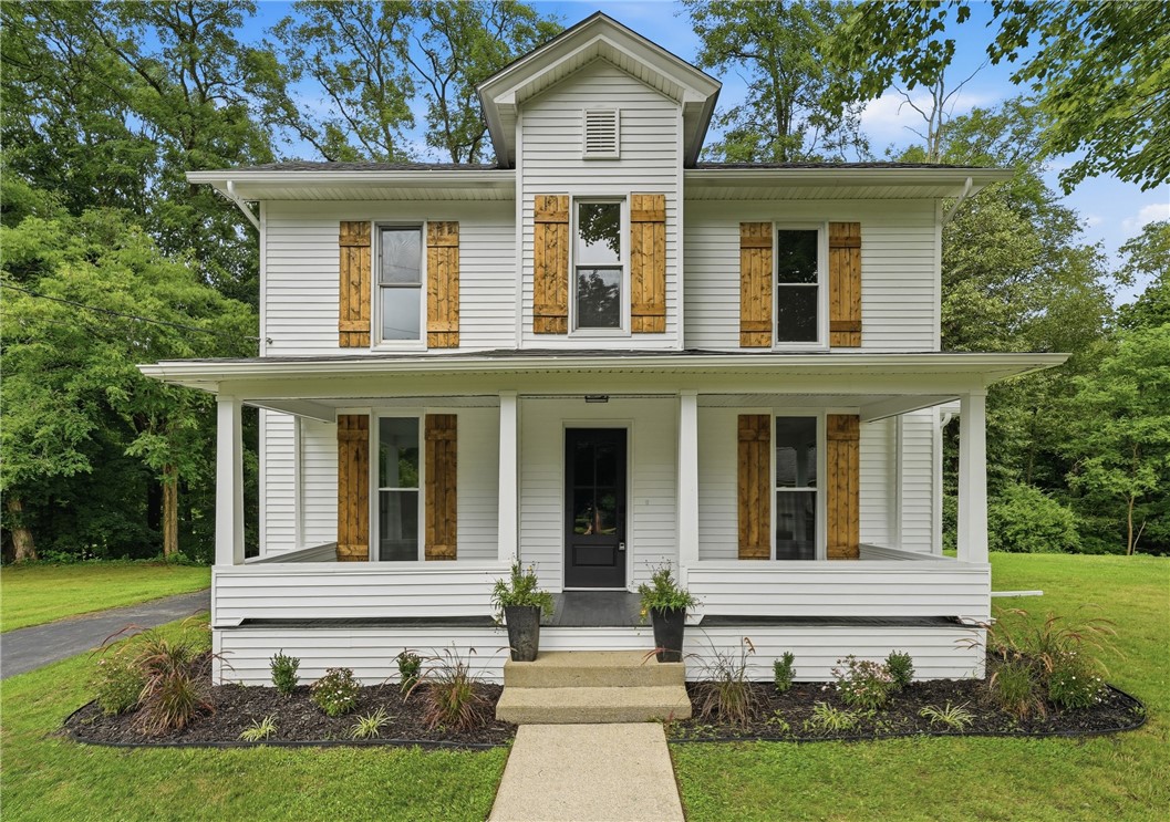 White two-story house with a wide covered porch, rustic wood shutters, and landscaped garden beds.