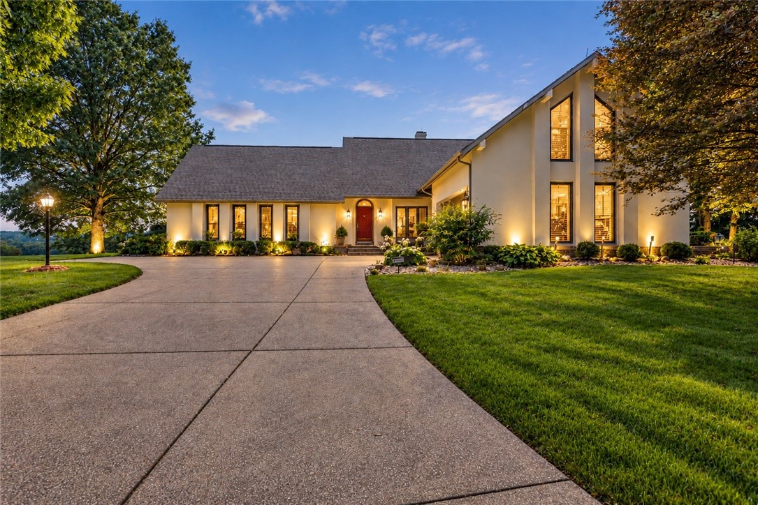 Large cream-colored home with a curved driveway, illuminated windows, and landscaped lawn at dusk.