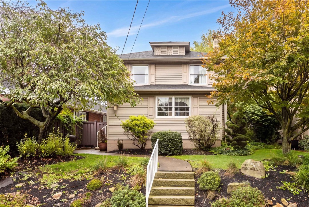 Beige two-story house framed by autumn trees, with a landscaped garden and central front steps.