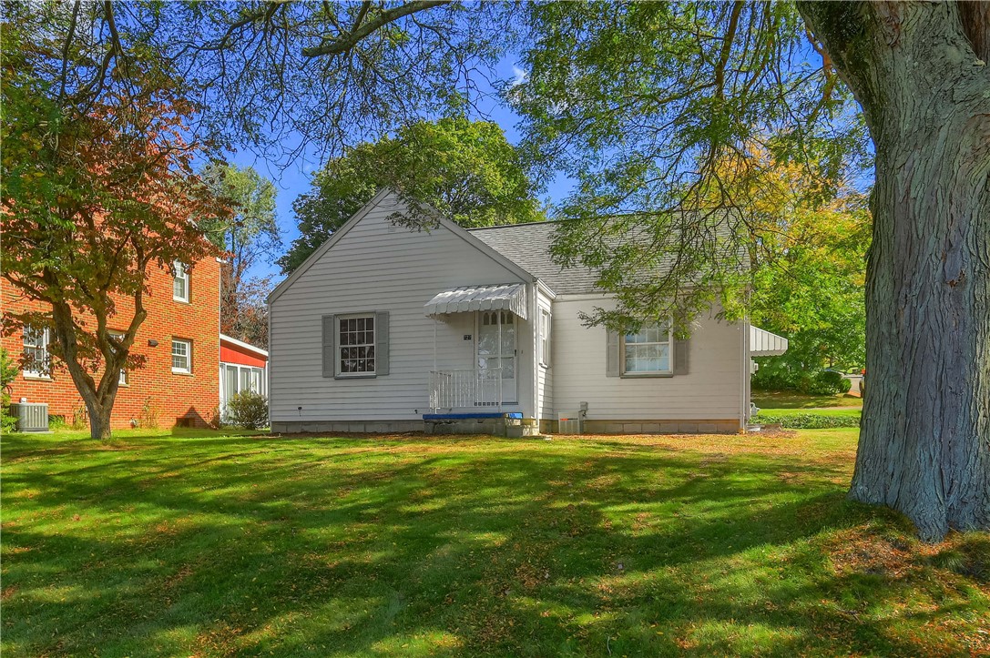 White-sided house with a small covered porch, shaded by large trees beside a brick building.