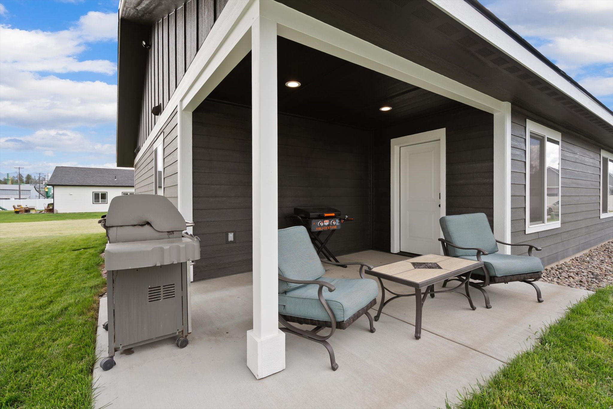 Covered patio with cushioned chairs, a table, and a grill beside a green lawn.