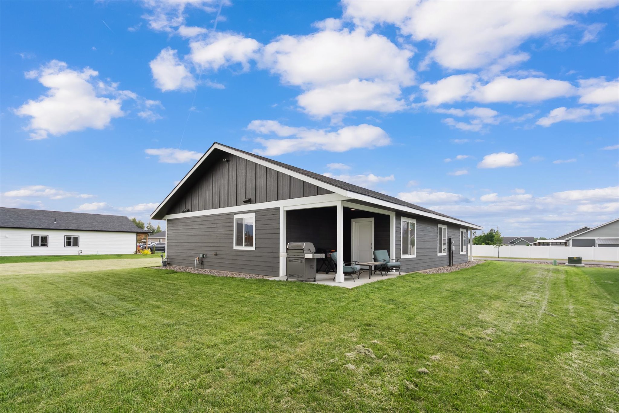 Gray single-story home with a covered patio, outdoor chairs, and grill beside a broad green lawn.