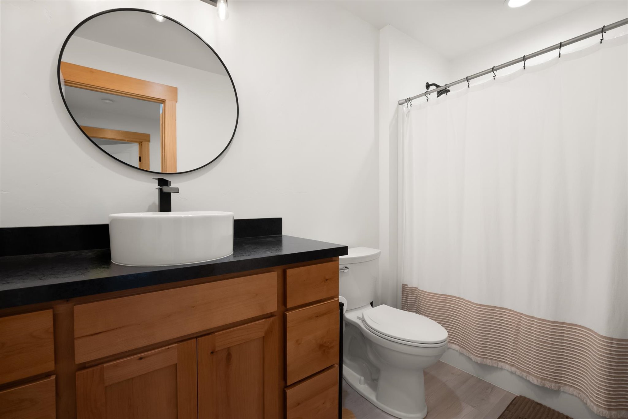 Bathroom with a wood vanity, round mirror, vessel sink, toilet, and striped shower curtain.