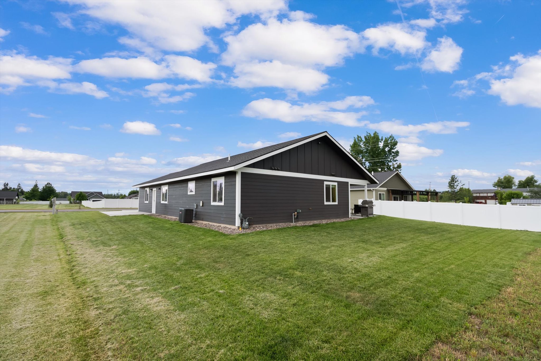 Dark gray single-story house with white trim, a broad lawn, and a white privacy fence.