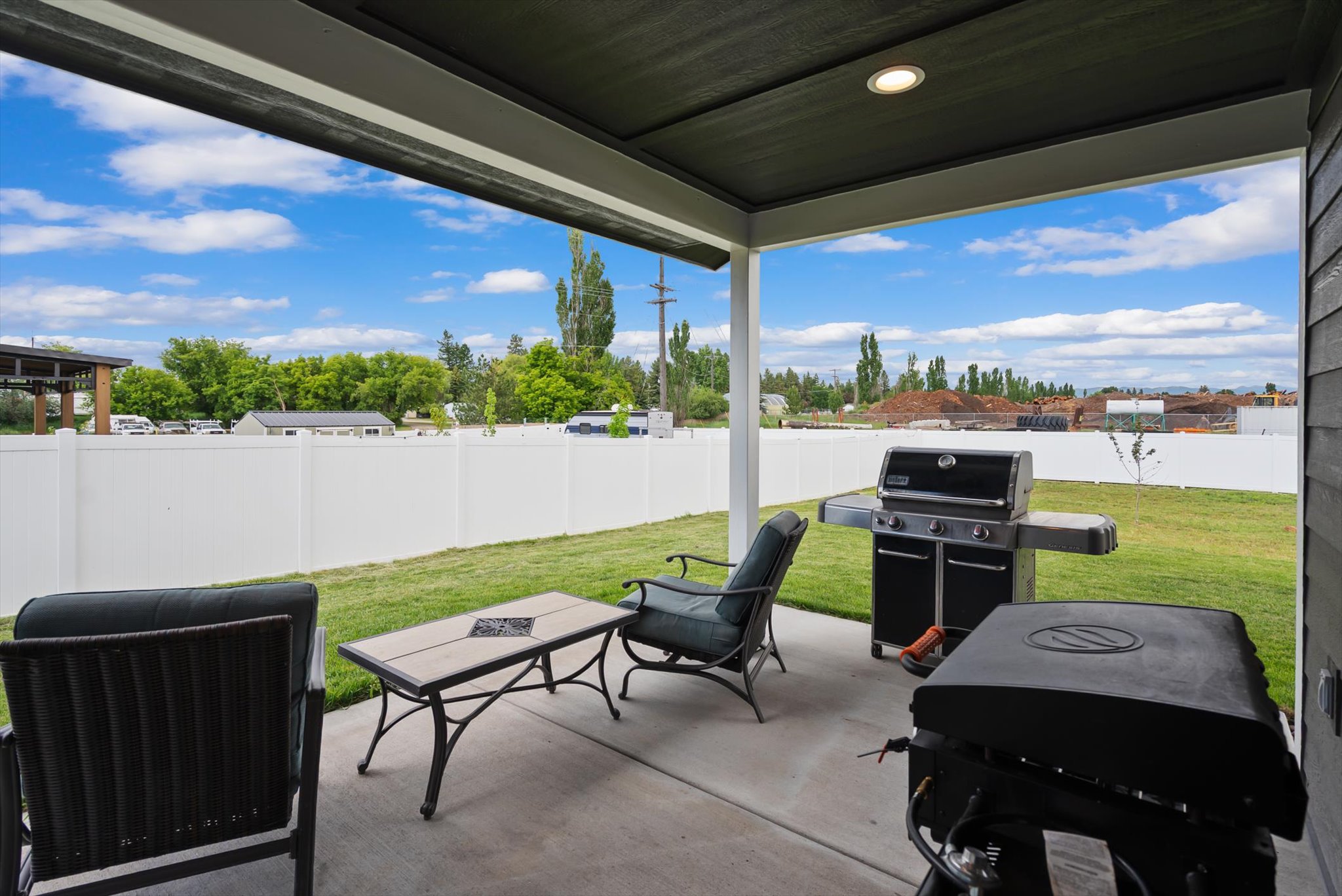 Covered backyard patio with outdoor seating, two grills, lawn, and white privacy fence.