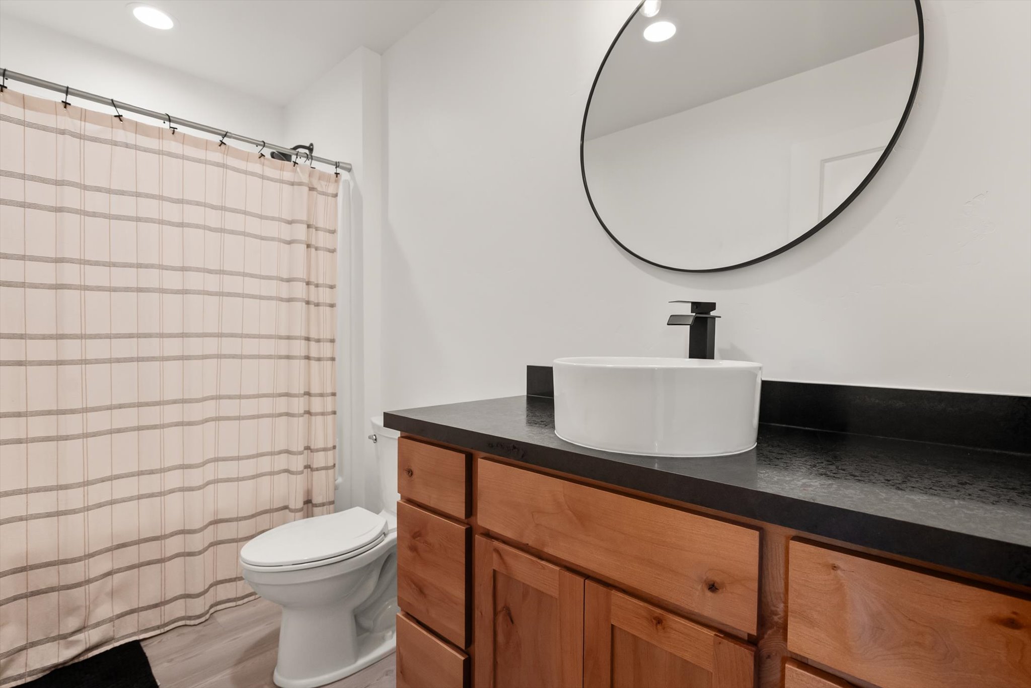 Bathroom with a wood vanity, vessel sink, and large round mirror beside a striped shower curtain.