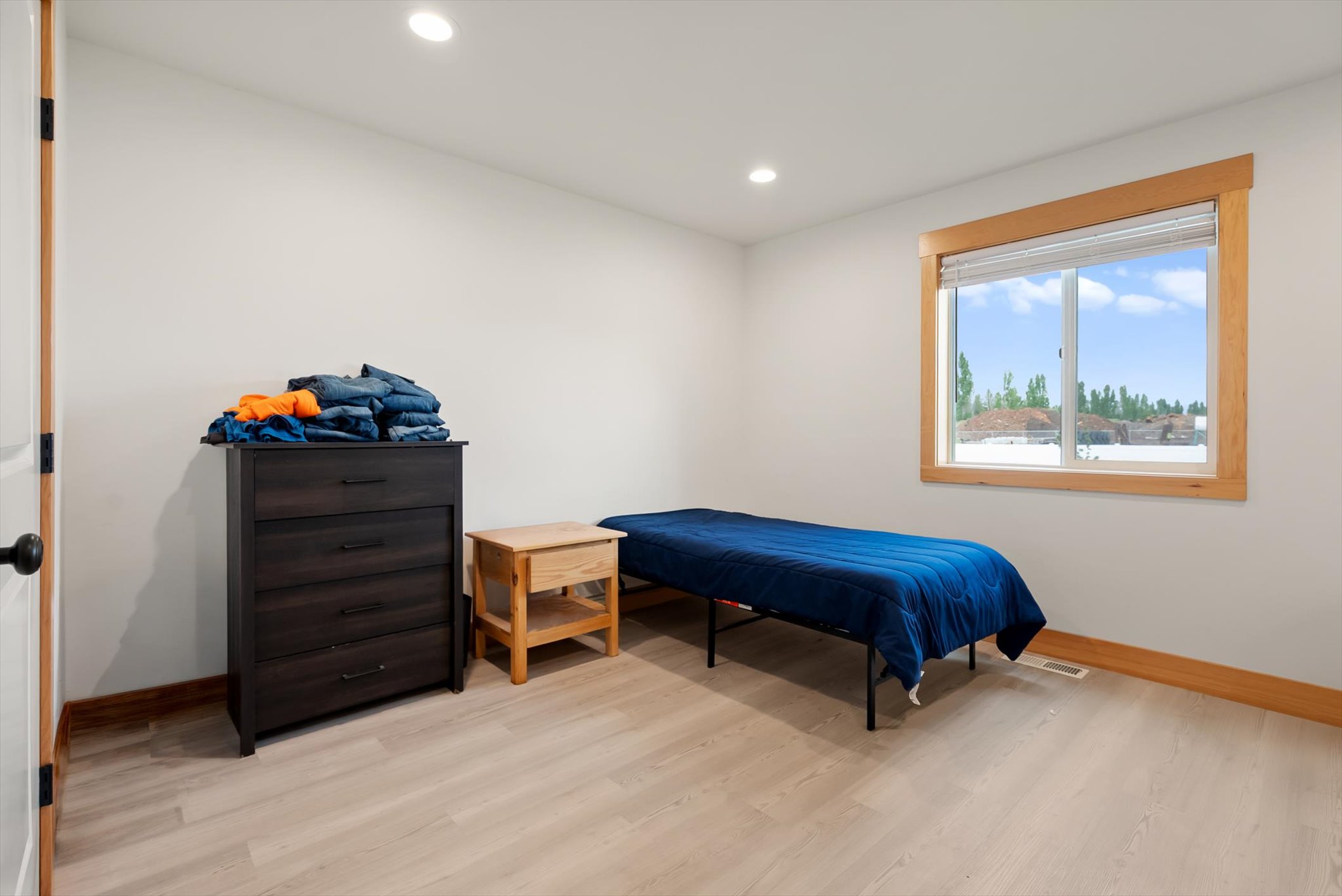 Bedroom with a navy-covered single bed, dark dresser, wooden nightstand, and wood-trimmed window.