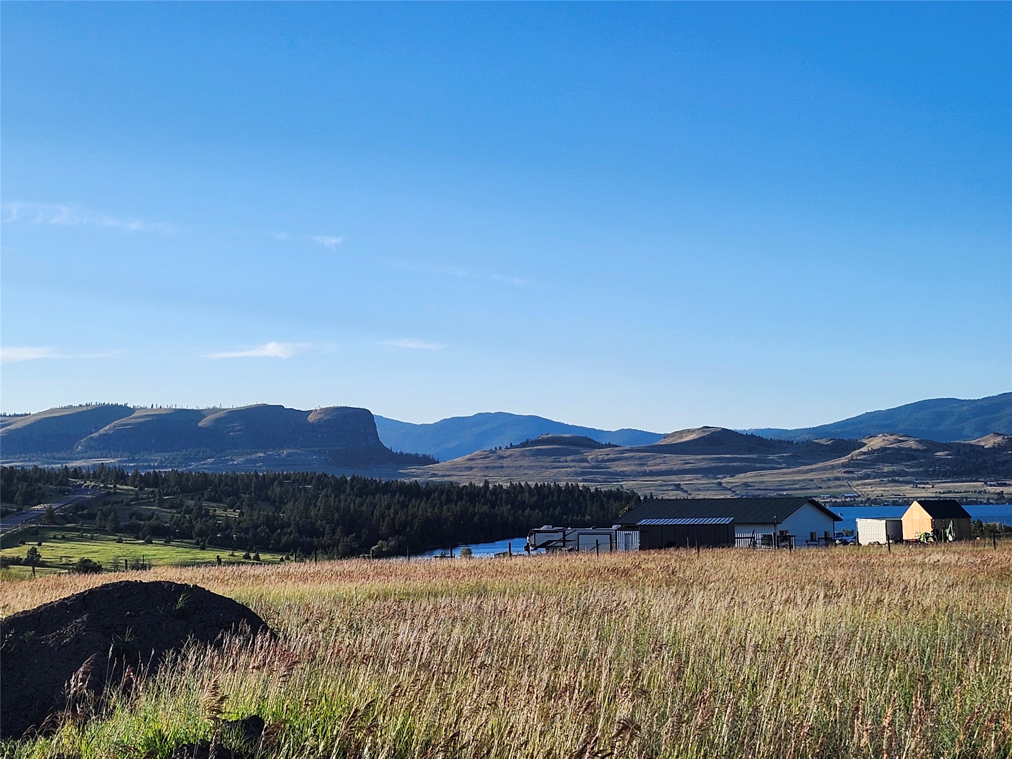 Rural property with outbuildings beside a lake, framed by golden grassland and mountain views.