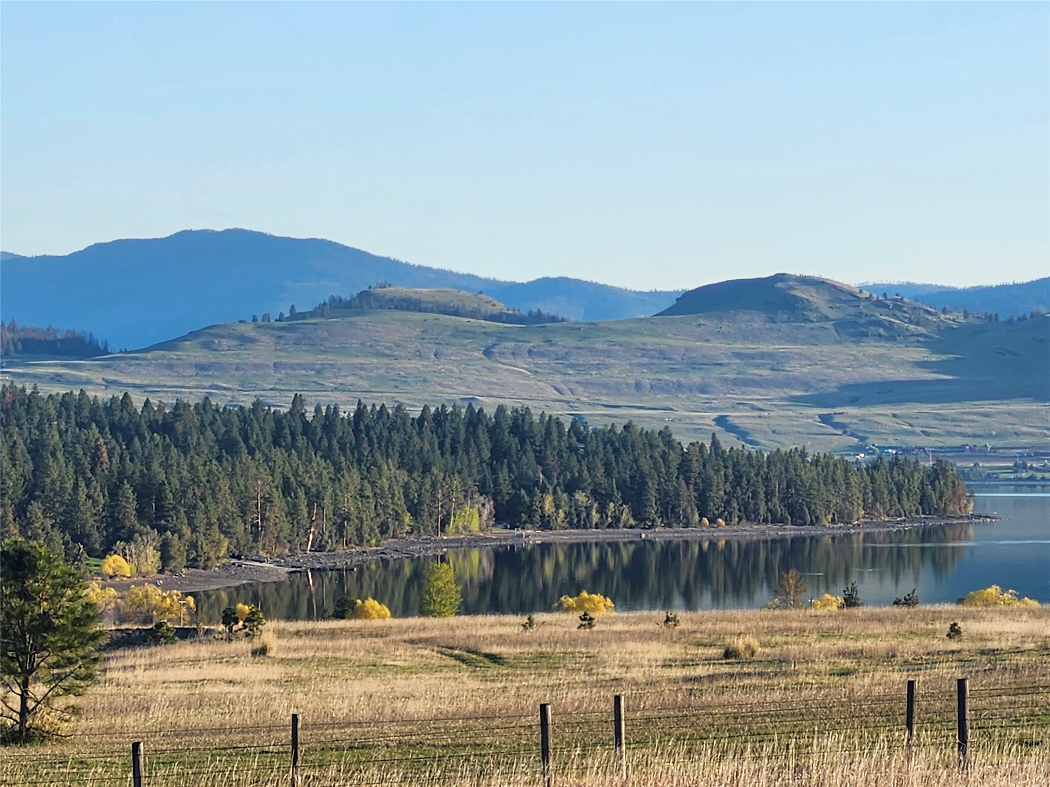 Lake and evergreen shoreline reflect below rolling mountains, viewed across a fenced grassy field.