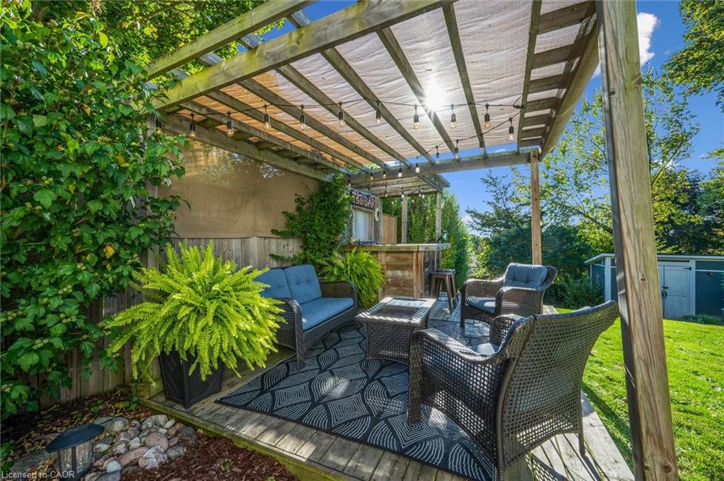 Backyard patio with wicker seating beneath a pergola, surrounded by ferns and leafy vines.