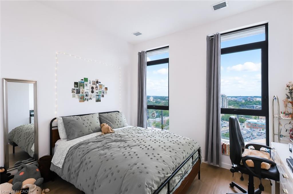 Furnished bedroom with a gray patterned bed, two tall city-view windows, and a desk with an office chair.
