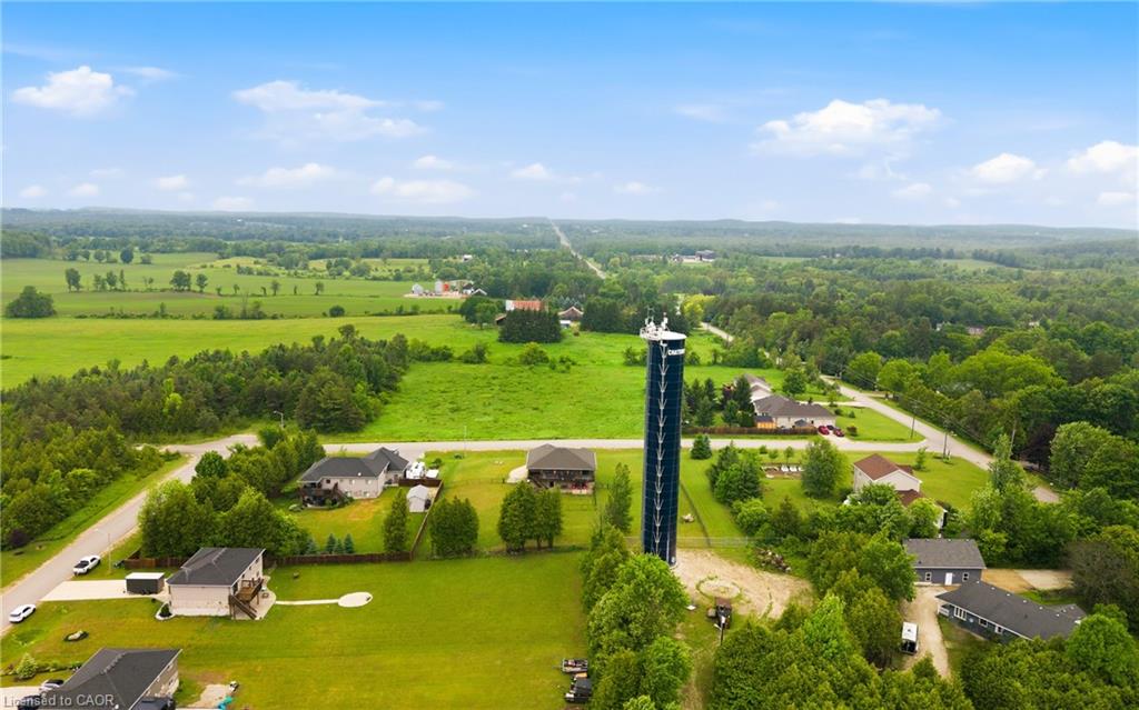 Aerial view of homes and green fields, with a tall dark tower and wooded countryside beyond.