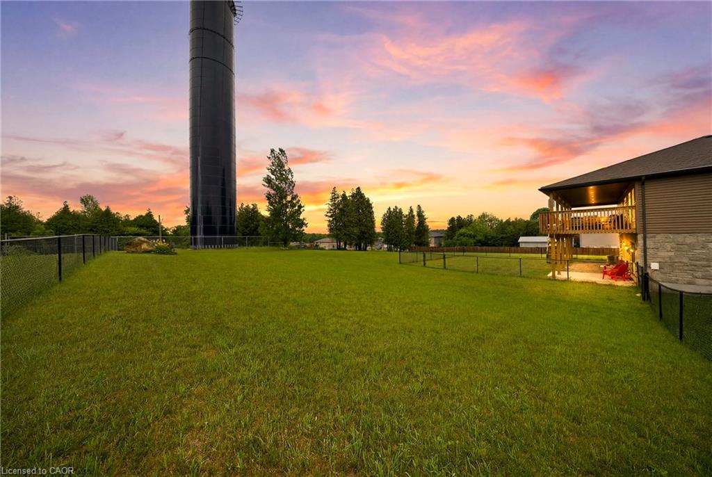 Grassy fenced backyard beside a house with a raised deck, water tower, and colorful sunset sky.
