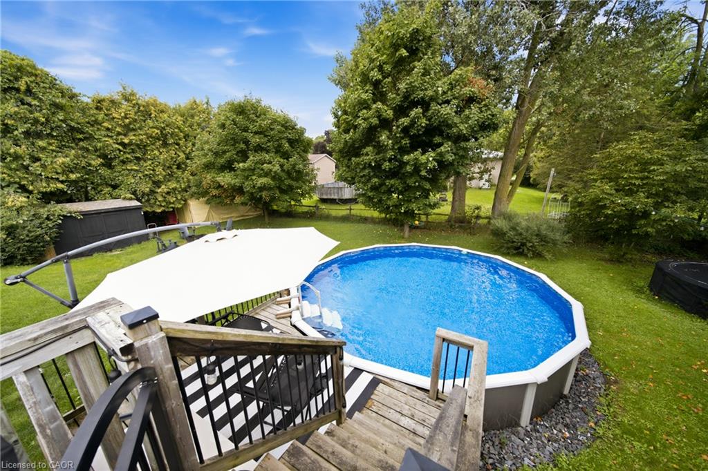 Above-ground pool beside a wooden deck, shaded by a large white umbrella in a leafy backyard.