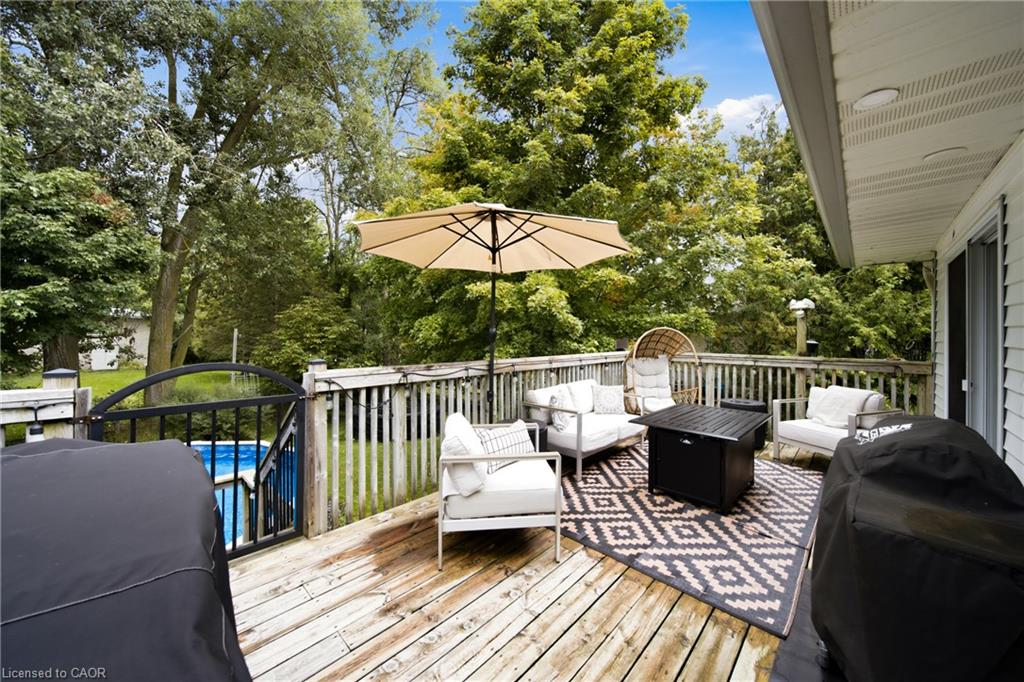 Wooden deck with white patio seating and umbrella beside a gated pool and leafy trees.