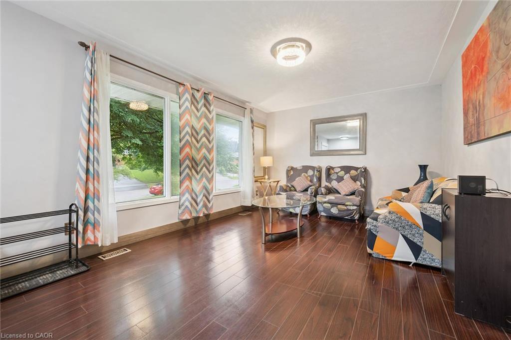 Furnished living room with dark wood floors, colorful curtains, and large windows overlooking a leafy street.