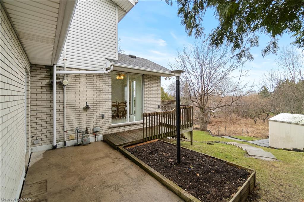 Rear patio beside a brick house, with a small deck, raised garden bed, and wooded yard.