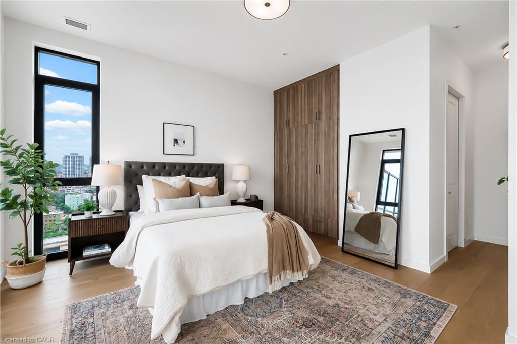 Staged bedroom with a white bed, wood wardrobe, and tall window overlooking the city.